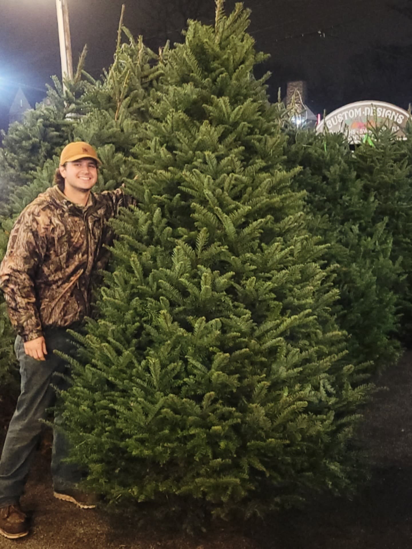 A Tree Guy beside a tall Noble fir at The Summit tree lot at night